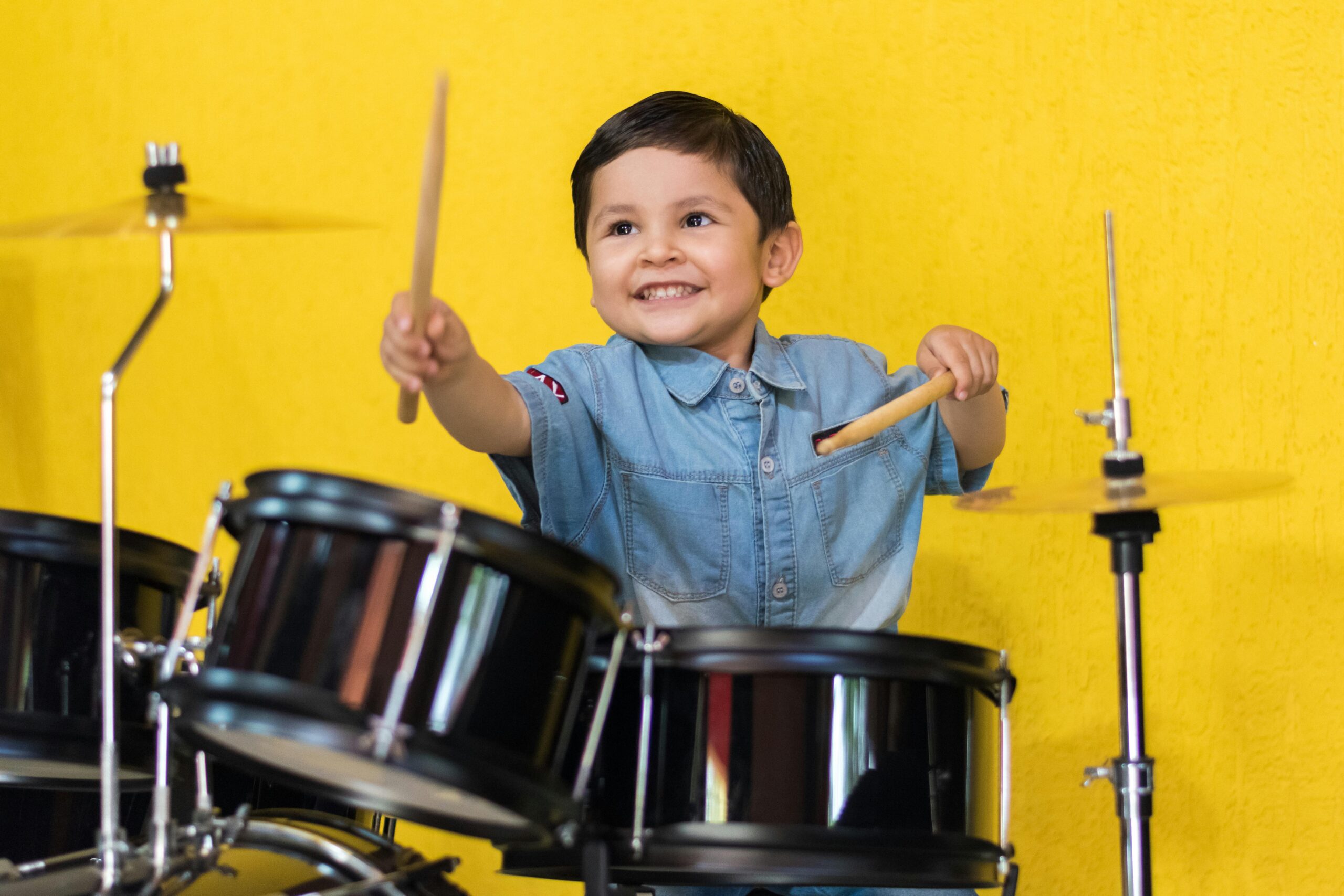 Young boy enthusiastically playing drums in front of a bright yellow wall, showcasing his musical talent.